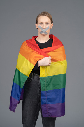 Person wrapped in a rainbow flag standing with their mouth taped