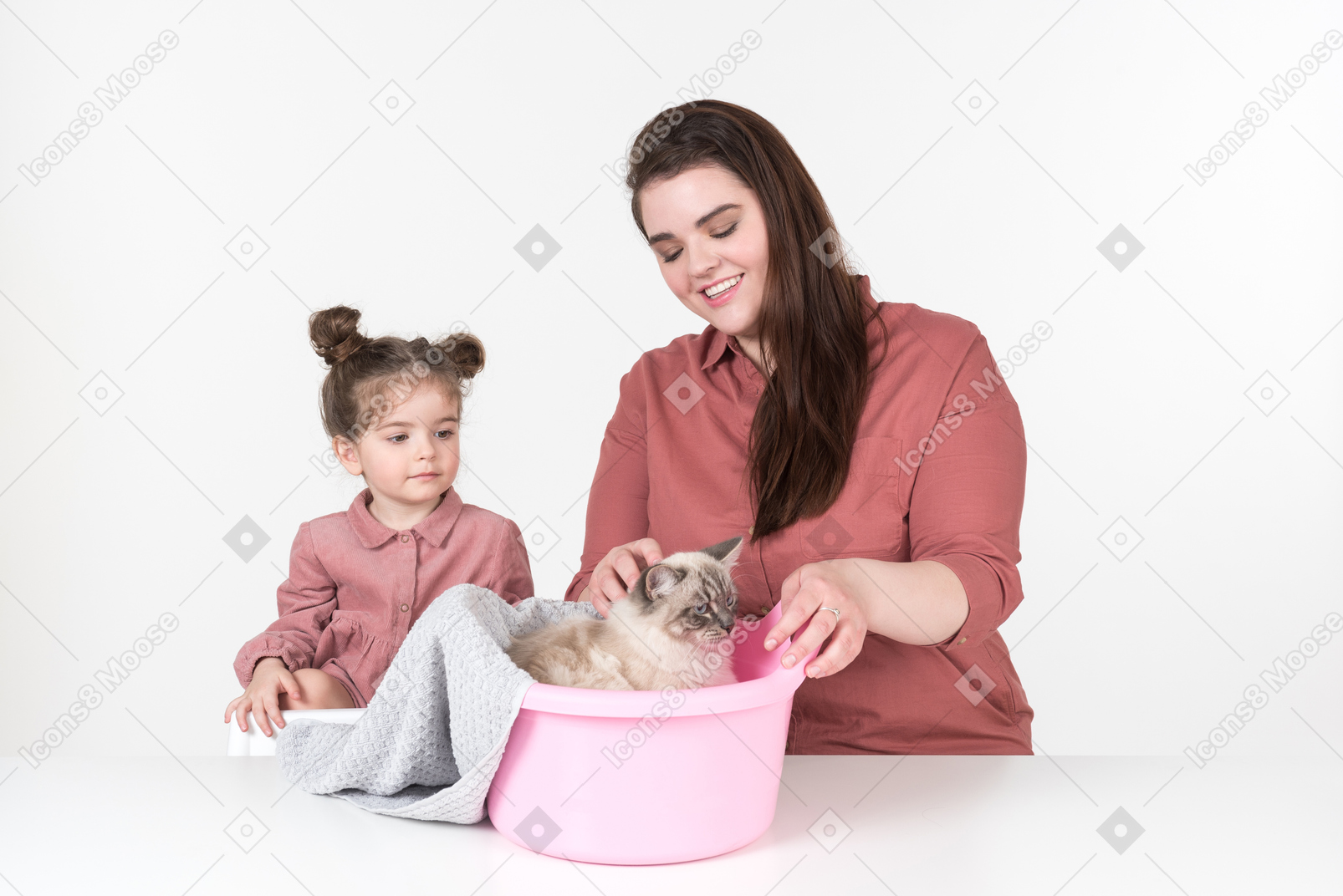 Madre y su pequeña hija, vestidas de rojo y rosa, sentadas a la mesa con su gato familiar