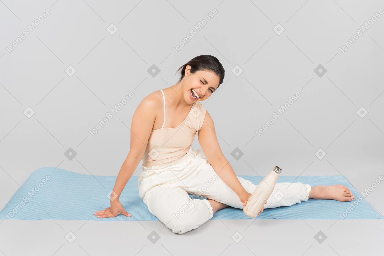 Lachende junge indische frau, die auf yogamatte sitzt und sportflasche hält