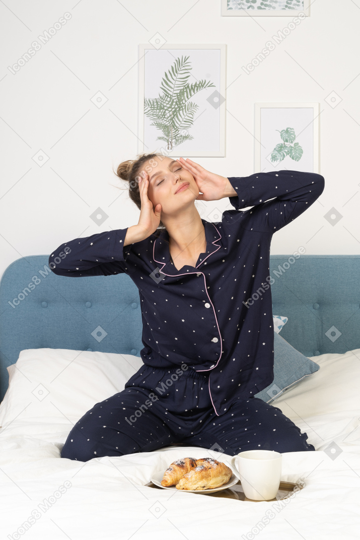 Front view of a pleased young lady in pajamas touching face before having breakfast in bed