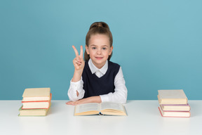 Kleines schulmädchen sitzt am schreibtisch und zeigt 'v' geste