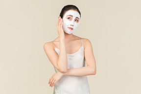 Rêveuse jeune femme asiatique avec masque facial, touchant sa main