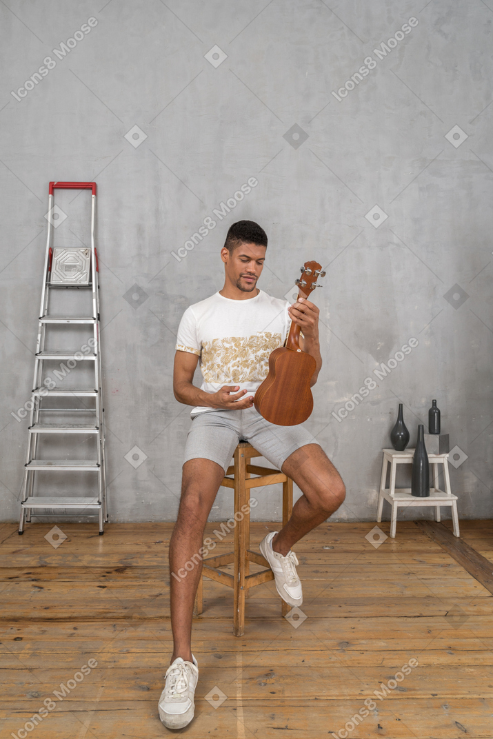 Vista frontal de um homem em um banquinho examinando um ukulele com um sorriso