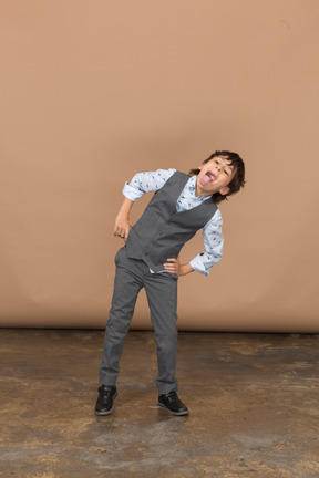 Вид спереди мальчика в костюме, позирующего с руками на бедрах и показывающего язык