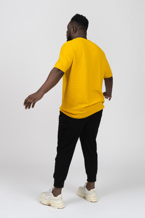노란 티셔츠를 입고 팔을 벌리고 있는 충격을 받은 검은 피부의 젊은 남자의 4분의 3 뒷모습