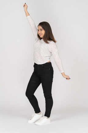 Vista de tres cuartos de una sonriente joven mujer india bailando en ropa casual levantando la mano