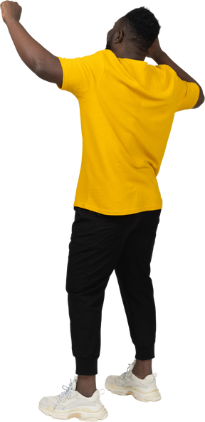 Three-quarter back view of a happy young dark-skinned man in yellow t-shirt raising hands