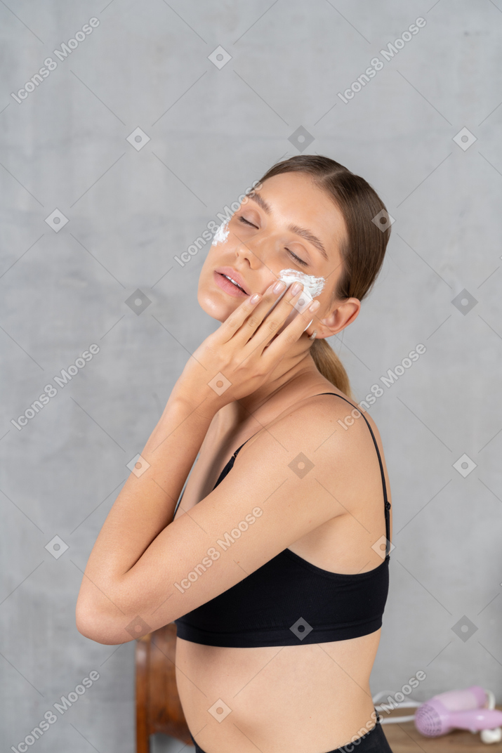 Mulher com olhos fechados aplicando creme facial nas maçãs do rosto
