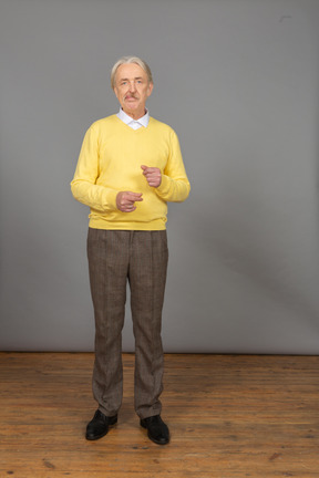 Front view of an old grimacing man in yellow pullover raising hands and looking at camera