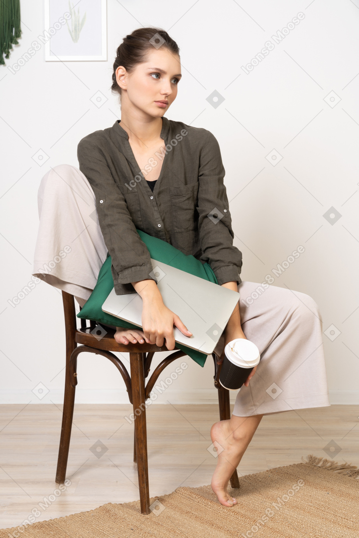 一位年轻女子坐在椅子上，拿着她的笔记本电脑和触摸咖啡杯的前视图