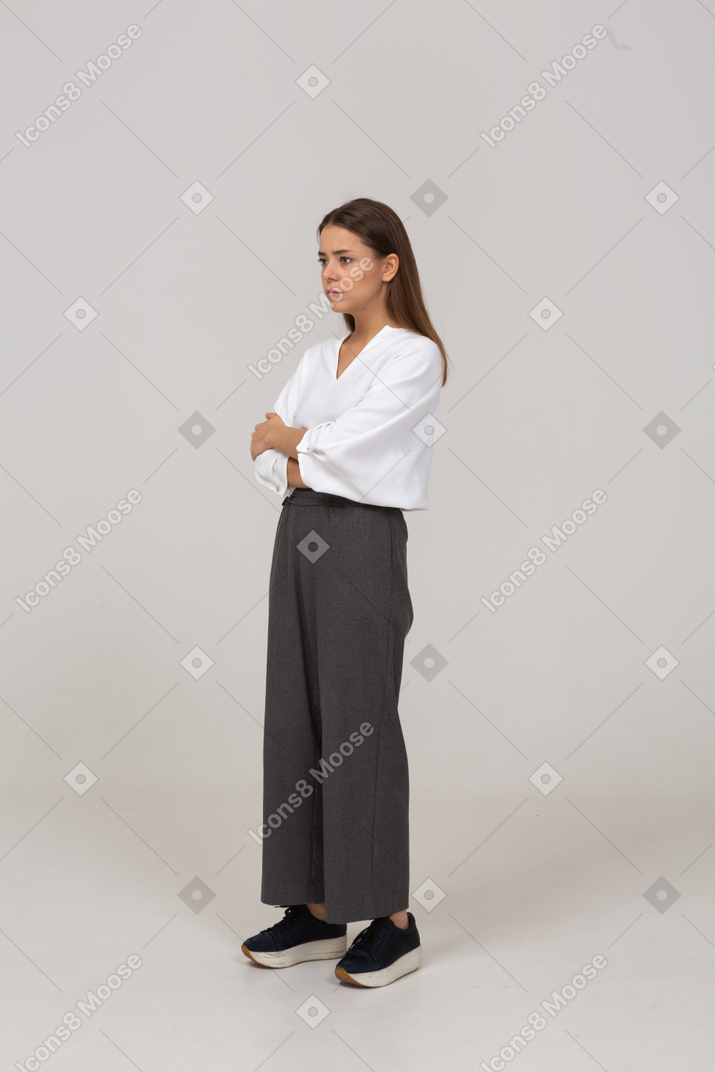 Вид в три четверти сомнительной молодой леди в офисной одежде, скрещивающей руки