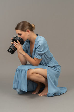 Three-quarter view of a young woman in blue dress sitting on a floor & taking shot