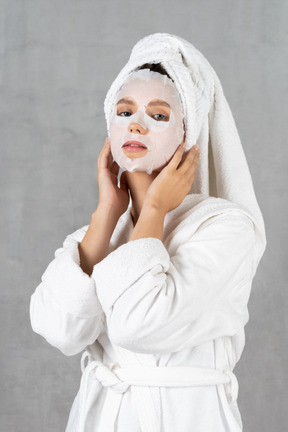 Woman in bathrobe with face mask on touching her face