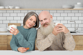 A man and woman sitting at a kitchen table holding coffee mugs
