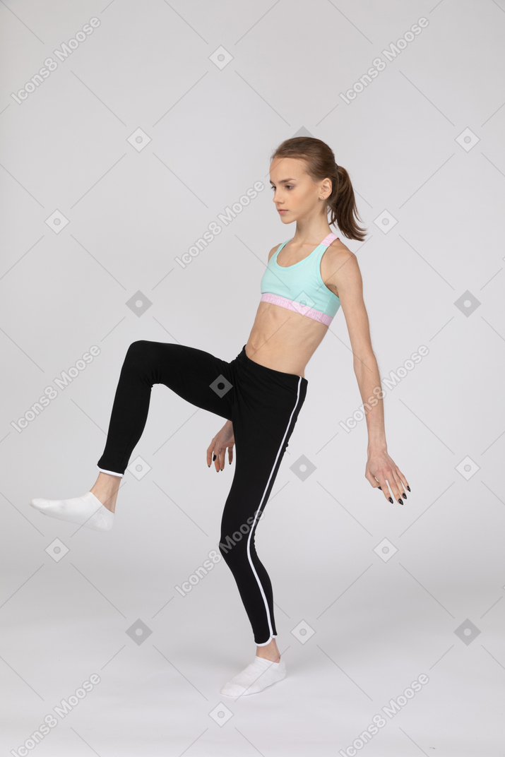 Three-quarter view of a teen girl in sportswear outspreading hands and raising leg
