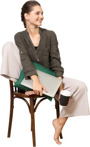 Front view of a smiling young woman sitting on a chair and holding her laptop & coffee cup