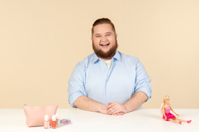 Rindo grande homem sentado à mesa com cosméticos e boneca barbie nele