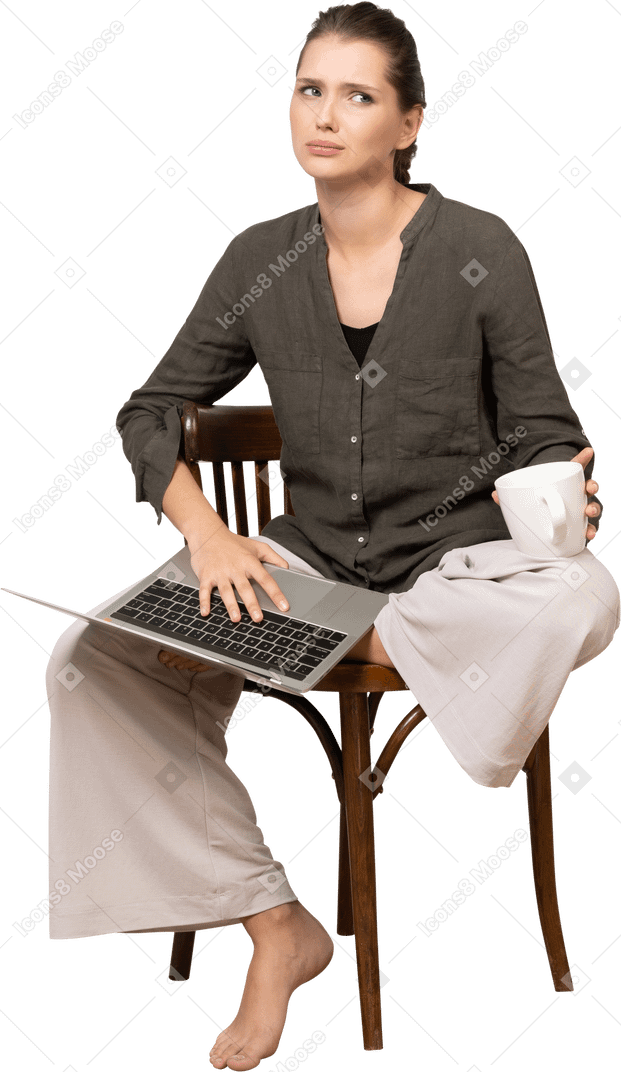 Vista frontal de una mujer joven confundida vistiendo ropa de casa sentado en una silla con un portátil y una taza de café
