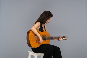 Vista de três quartos de uma jovem sentada de terno preto tocando violão e olhando para baixo