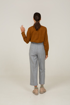 Vista traseira de uma jovem mulher asiática de calça e blusa, mostrando um gesto de ok