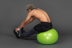 Vista posterior de tres cuartos de un hombre afro sin camisa que se extiende mientras está sentado en una pelota de gimnasia verde