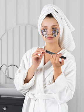 A young woman in a robe with eye patches filing her nails in a bathroom