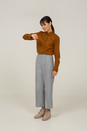 Vista di tre quarti di una giovane donna asiatica in calzoni e camicetta che mostra il pollice verso il basso