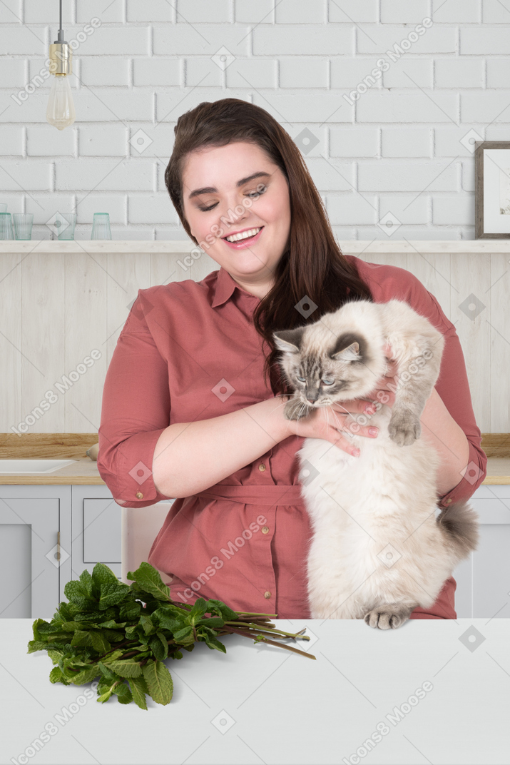 A plus-size woman holding a cat near a bunch of catnip lying on the table in front of them