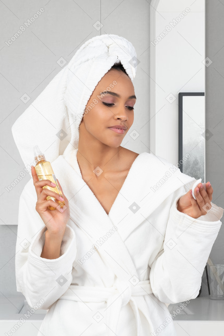 A woman in a bathrobe holding a spray bottle and a cotton pad