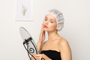 Young female patient in medical cap touching her skin while looking in the mirror