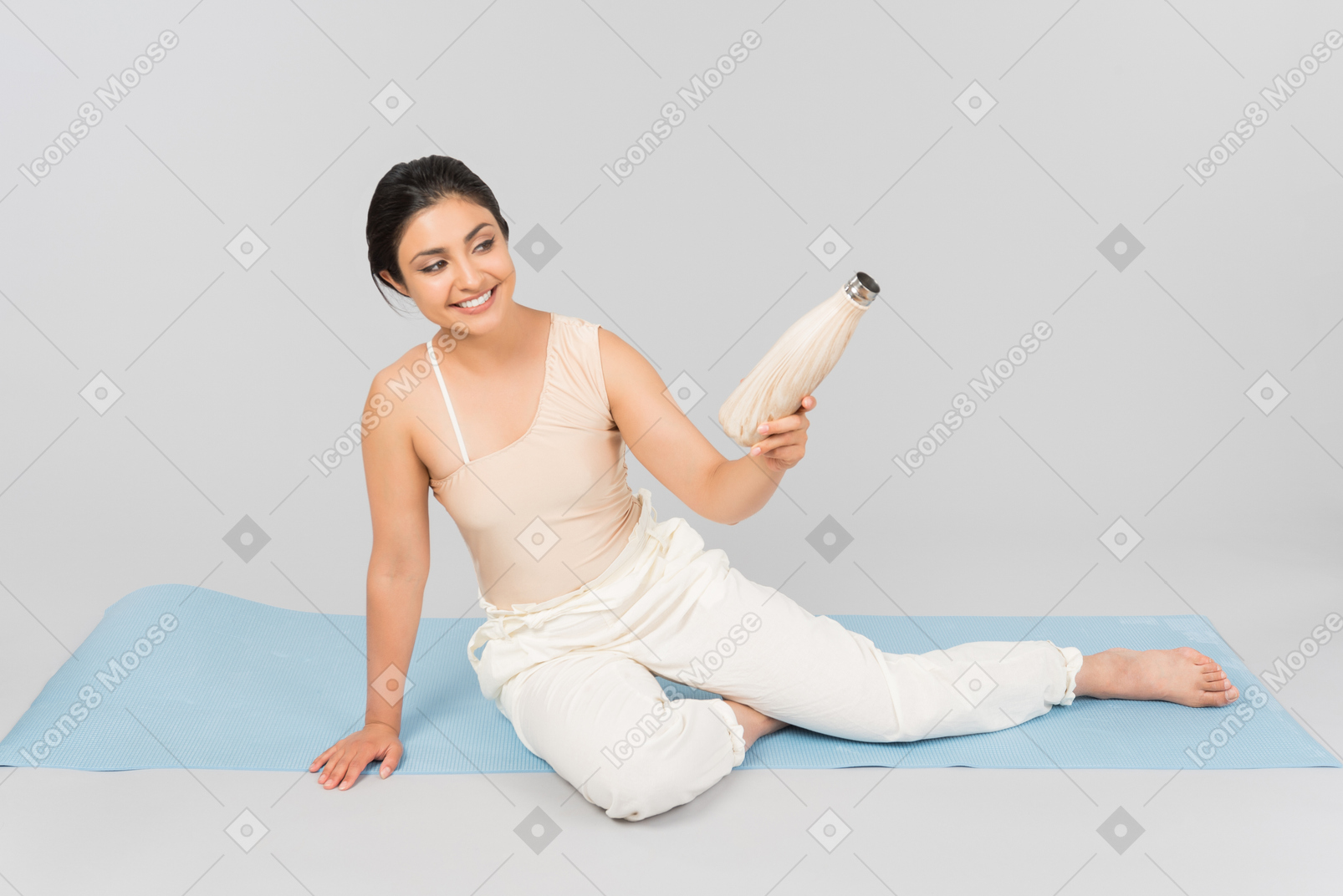 年轻的印度女人坐在瑜伽垫上，拿着运动水壶
