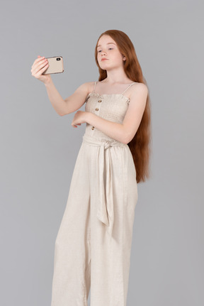 Teenage girl dressed in beige overalls making a selfie with smartphone