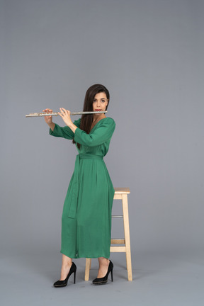 Девушка в зеленом платье в полный рост сидит на стуле и играет на кларнете