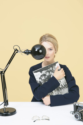 Young woman holding a magazine and sitting at the table with lamp and camera on it