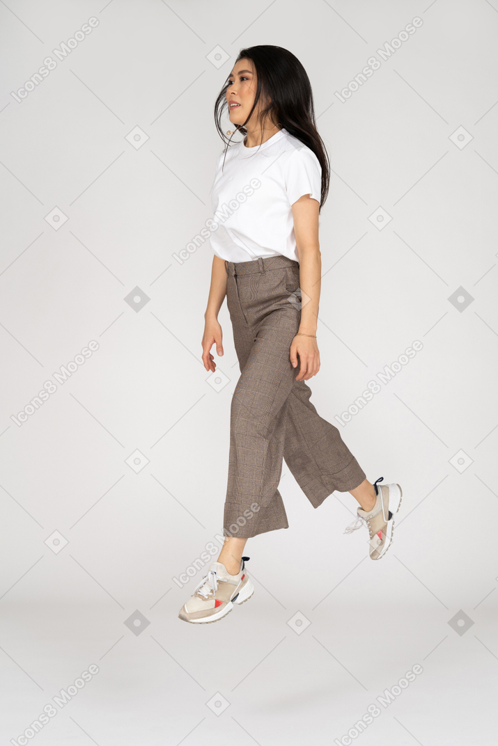 Vista di tre quarti di una giovane donna che salta in calzoni e maglietta che allarga le gambe