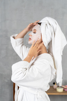 Side view of a woman in bathrobe applying a sheet mask