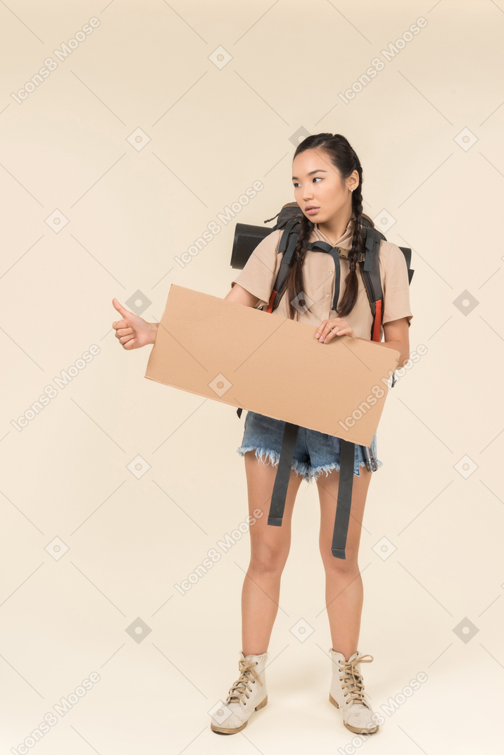 Junge weibliche anhalterin, die papierkarte hält und sich daumen zeigt
