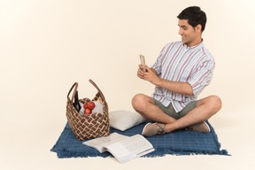 Young caucasian guy sitting on blanket and making a photo of picnic basket