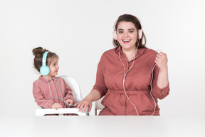 Mother and her little daughter, dressed in red and pink clothes, sitting at the dinner table, listening to the music together