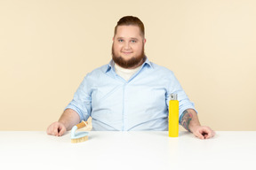 Contented looking young overweight househusband sitting at the table with cleaning spray and brush on it