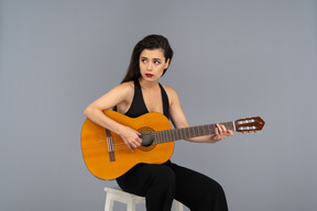 Front view of a sitting young lady in black suit playing guitar and looking aside