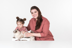 La mamma e la figlia piccola, vestite con abiti rossi e rosa, sedute a tavola con il loro gatto di famiglia