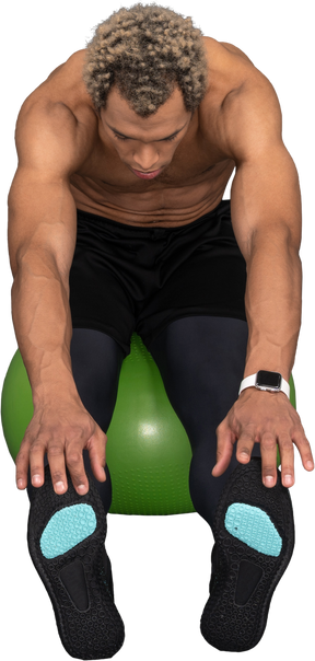 Front view of a shirtless afro man stretching while sitting on a green gym ball