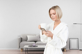 A woman in a bathrobe holding a cup of coffee and looking at a cell phone