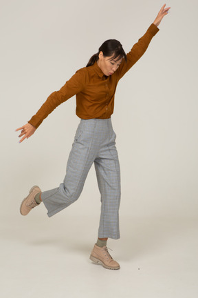 Vista de três quartos de uma jovem mulher asiática de calça e blusa, levantando a mão e levantando a perna