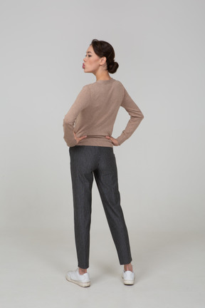 Vista posteriore di tre quarti di una giovane donna in pullover e pantaloni che fa il broncio e mette le mani sui fianchi