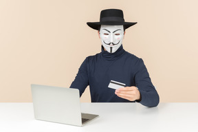 Hacker wearing vendetta mask sitting at the laptop and holding bak card