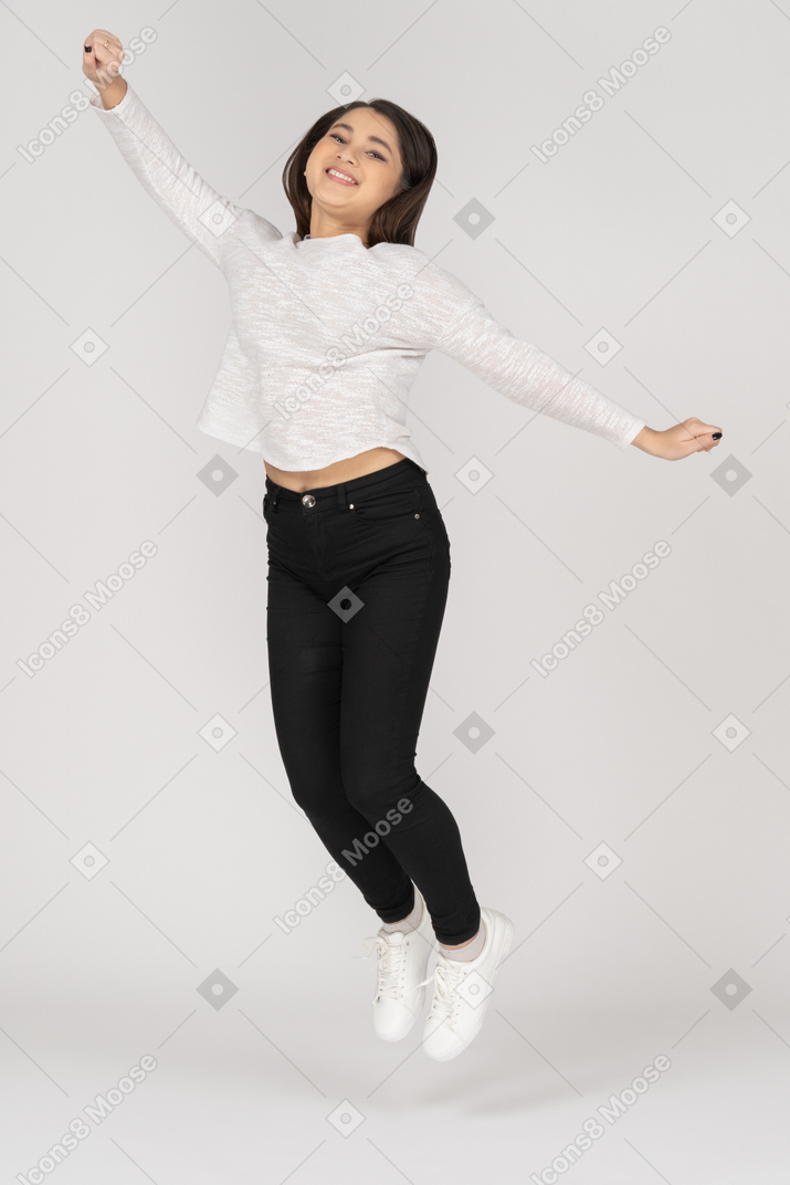 Vista de três quartos de uma jovem índia sorridente e pulando em roupas casuais, levantando a mão