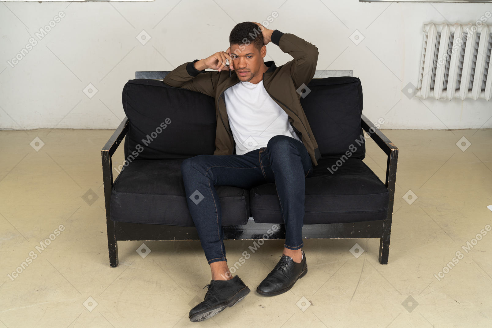 Вид сбитого с толку молодого человека, сидящего на диване и разговаривающего по телефону