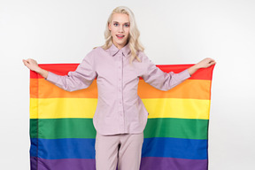 Young blond-haired person in pastel pjs and a rainbow flag in their hands, standing against the plain white background